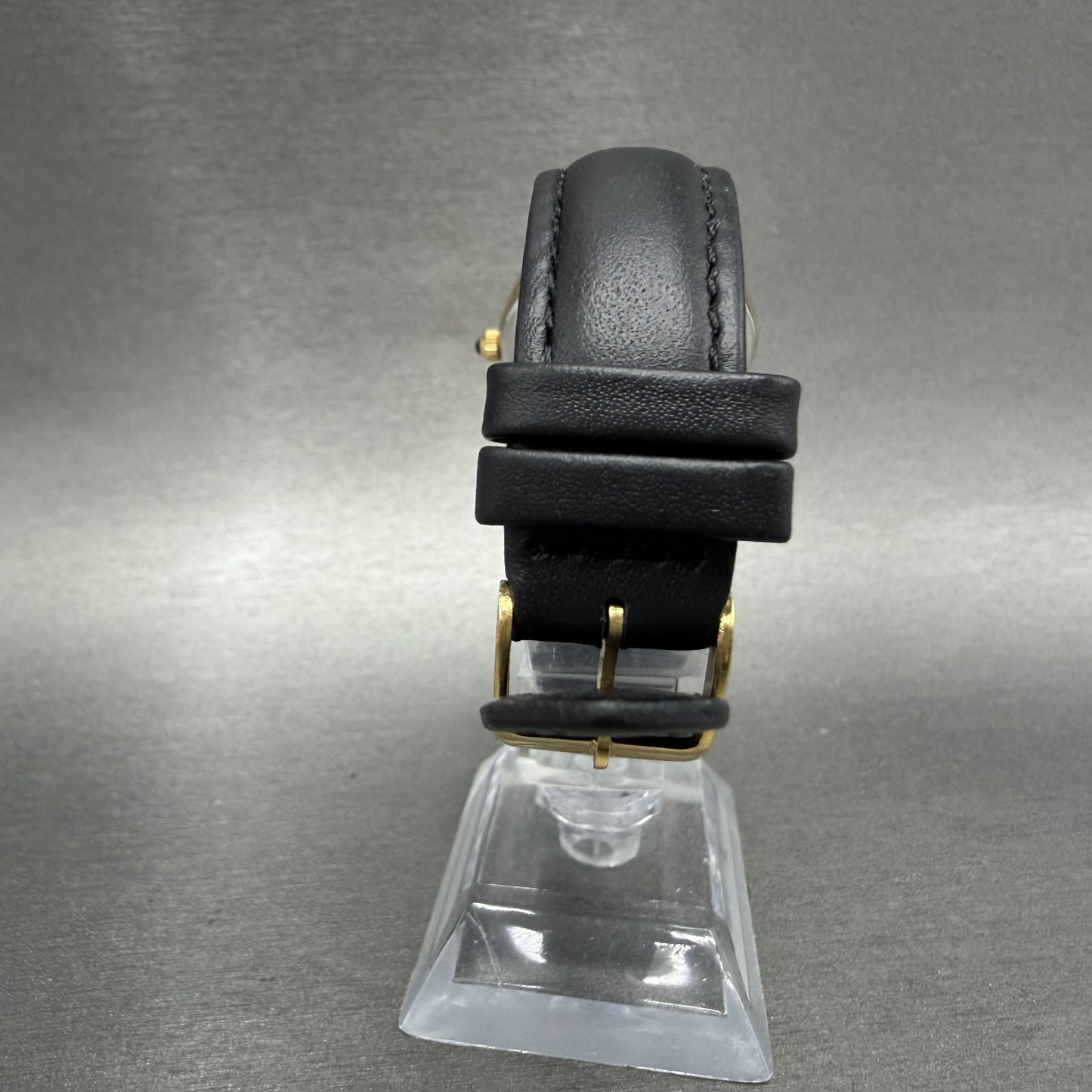 Black leather watch strap with gold buckle on display stand against grey background