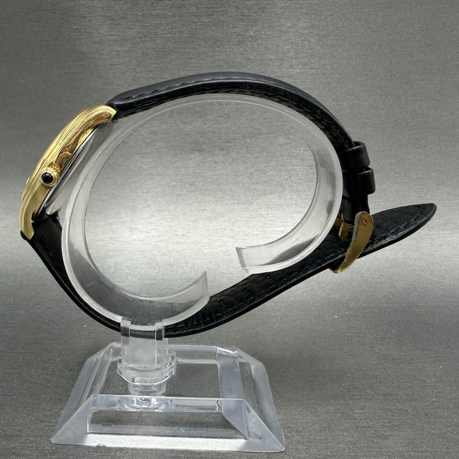 Gold wristwatch with black leather strap on clear display stand, gray background