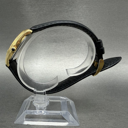 Gold wristwatch with black leather strap on clear display stand, gray background