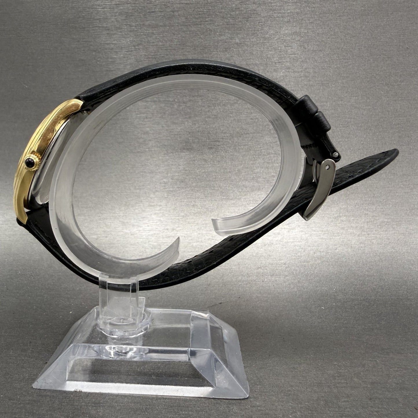 Gold dress watch with black leather strap on clear display stand, grey background