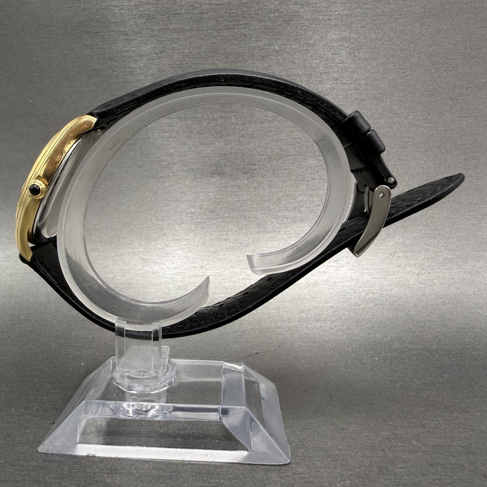 Gold dress watch with black leather strap on clear display stand, grey background